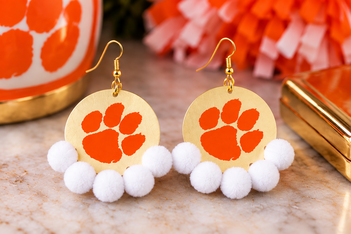 Pair of Clemson paw statement earrings with orange paw designs, gold-tone round pendants, hook closures and white pom-pom trim.