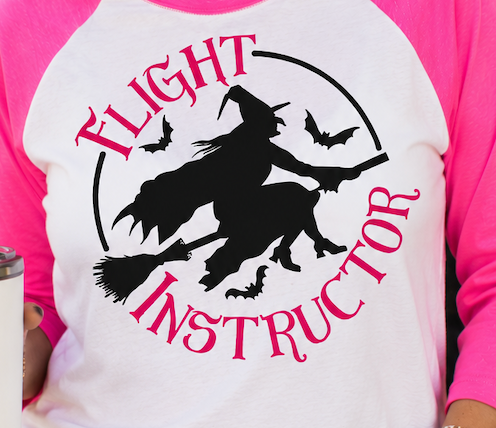 A close-up of a person wearing a white and bright pink raglan t-shirt with a black and pink graphic that reads 'FLIGHT INSTRUCTOR' around a silhouette of a witch riding a broomstick with bats.