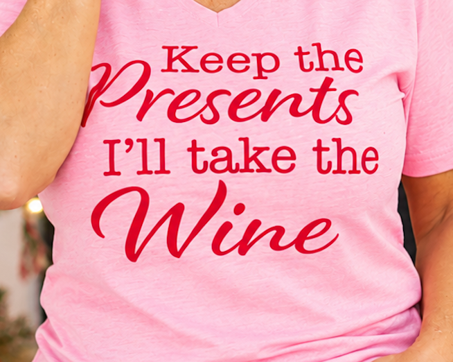 Close-up of red Keep the Presents, I’ll Take the Wine lettering on a pink V-neck Christmas tee