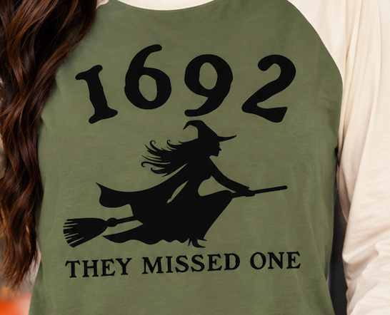 Close-up of the black 1692 witch design and “They Missed One” text on the military green body of the cream-sleeve raglan.