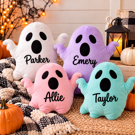 Four plush ghost pillows in white, purple, pink, and teal are arranged on a woven rug with fall decor, including pumpkins, a lantern, and string lights. Each pillow has a black name printed on it: Parker, Emery, Allie, and Taylor.