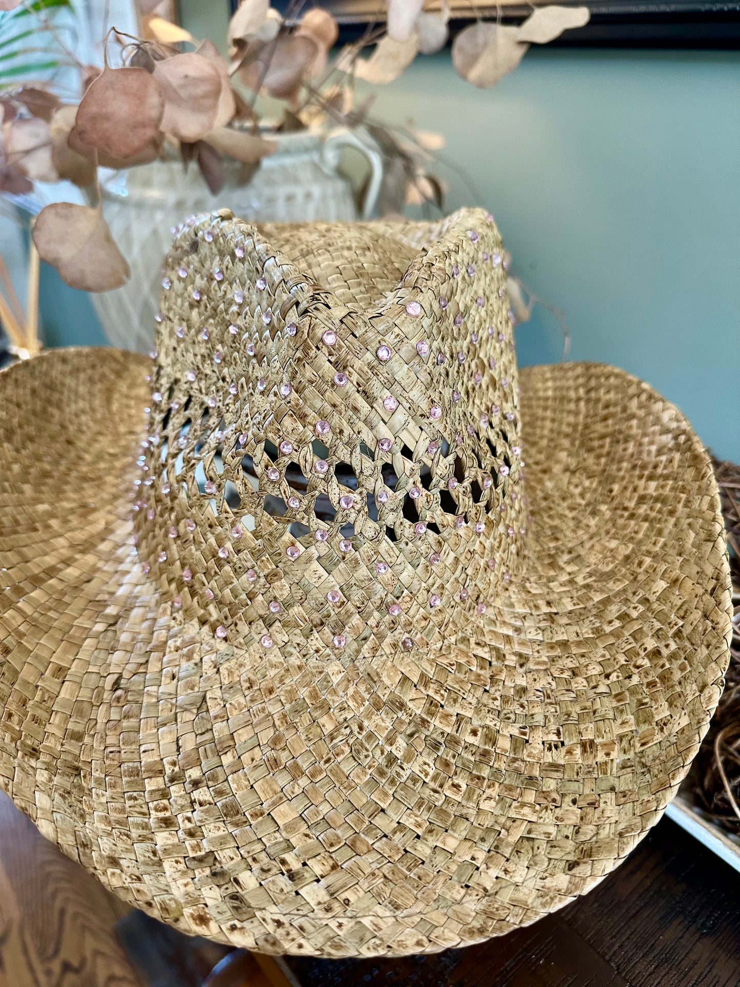 A woven straw cowboy hat with a wide brim and a textured crown, embellished with pink rhinestones, resting on a dark wooden surface.