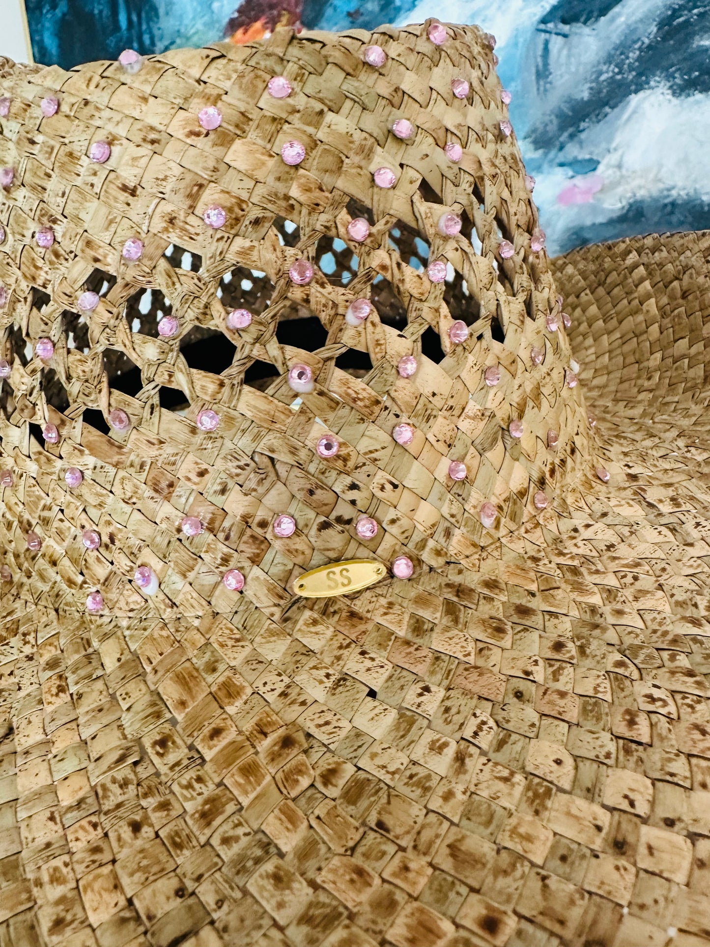 A close-up of a woven straw cowboy hat decorated with pink rhinestones and a small gold metal badge on the crown, set against a colorful painted backdrop.