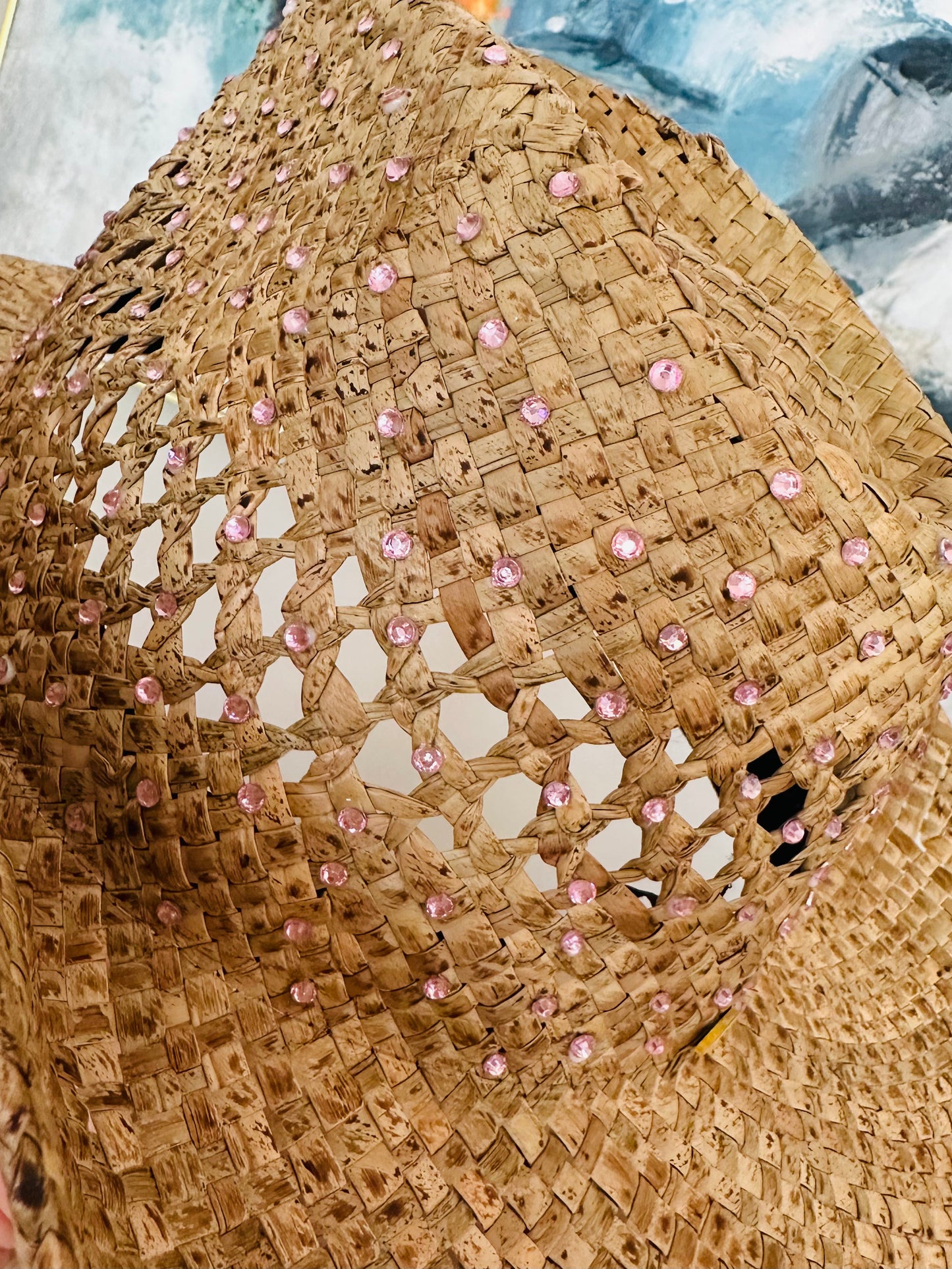 A close-up of a woven straw hat with a wide brim, decorated with small pink rhinestones set into the weave.