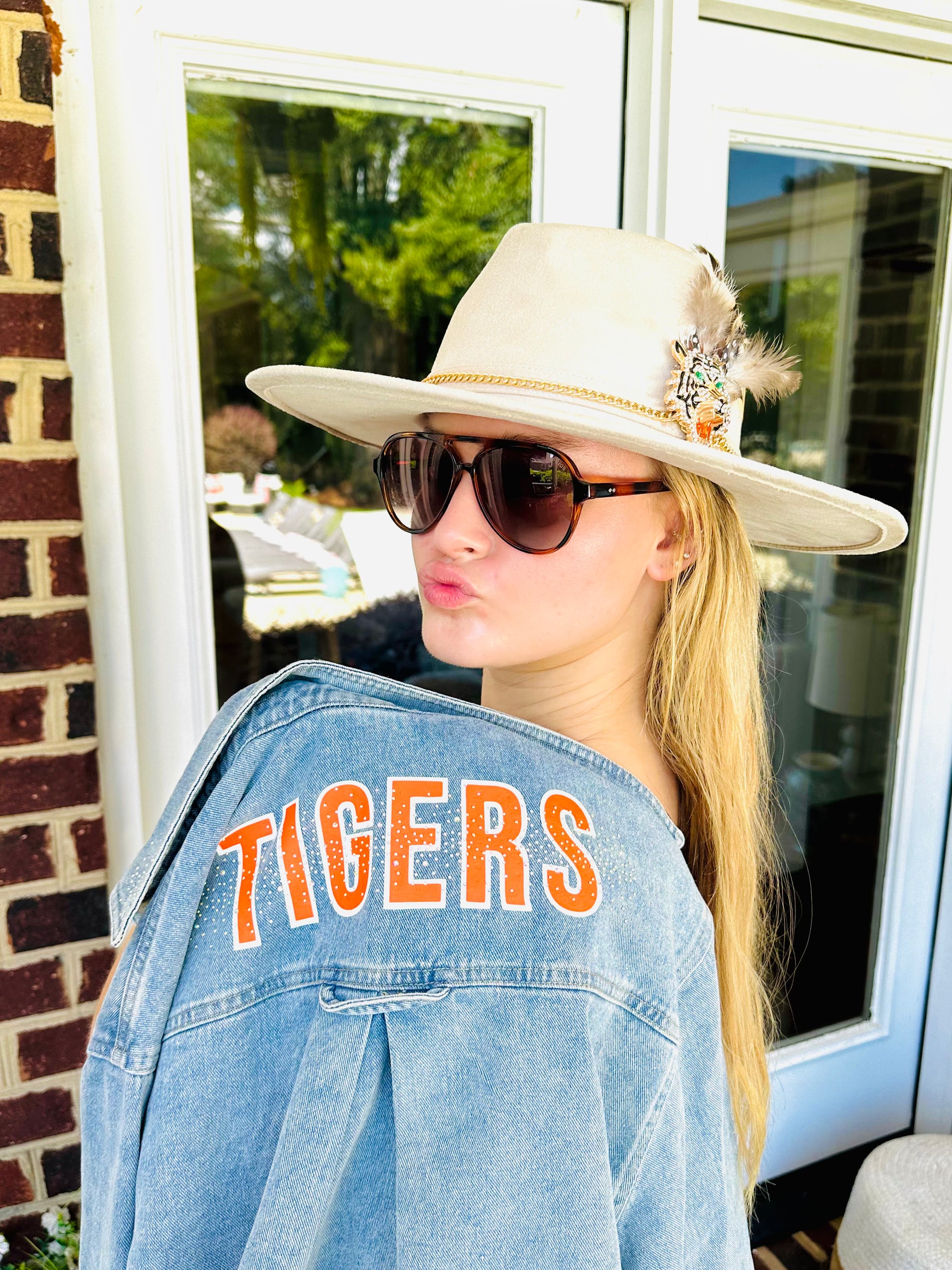 Woman wearing a cream Clemson Show Stopping Topper with jeweled tiger, gold chain and feather details styled with a TIGERS denim jacket.