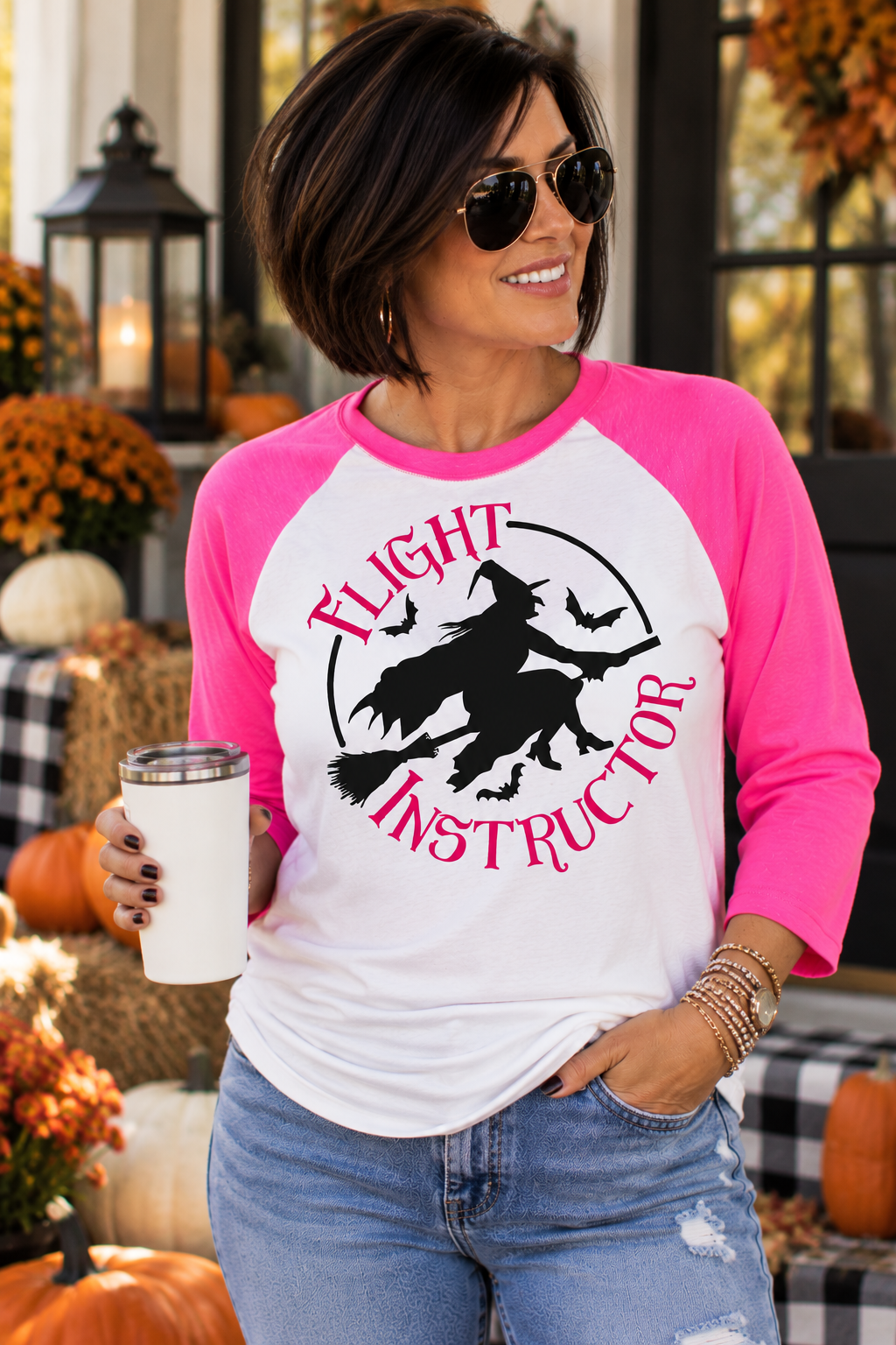 A woman wearing a pink and white raglan baseball t-shirt with a black graphic of a witch riding a broomstick and the words 'Flight Instructor'. She is holding a white travel tumbler in a fall-themed outdoor setting with pumpkins and autumn decorations.