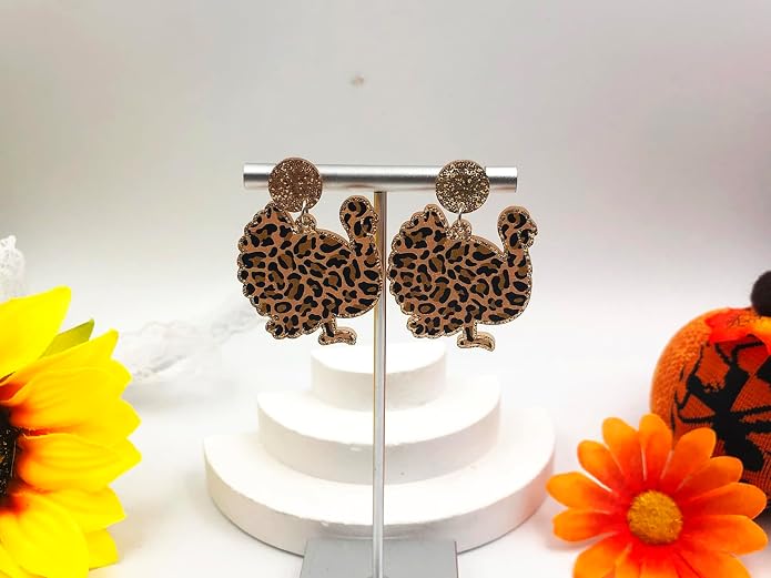 Pair of leopard-print turkey dangle earrings with a textured gold-tone top displayed on a stand beside fall flowers and a pumpkin.
