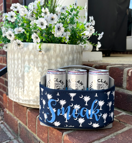 A navy blue can cooler with a white palmetto tree and moon pattern and the word 'Smoak' in light blue script, holding three Clam Creek beer cans, set on a brick patio next to a potted plant.